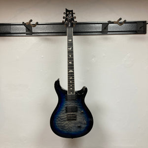 A PRS SE Mark Holcomb - Holcomb Blue Burst w/Gigbag Used Consign hangs on a black wall-mounted holder against a white wall.
