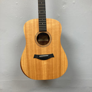 Taylor Academy 10e Acoustic-Electric Guitar w/GigBag in natural wood finish hangs on a gray wall, displaying the body and part of the neck.