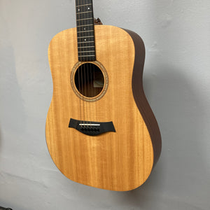 A Taylor Academy 10e Acoustic-Electric Guitar w/GigBag in light brown with six strings hangs on a light gray wall.