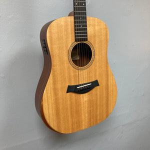 A Taylor Academy 10e Acoustic-Electric Guitar w/GigBag with a light brown body and dark fretboard hangs against a plain light gray wall.