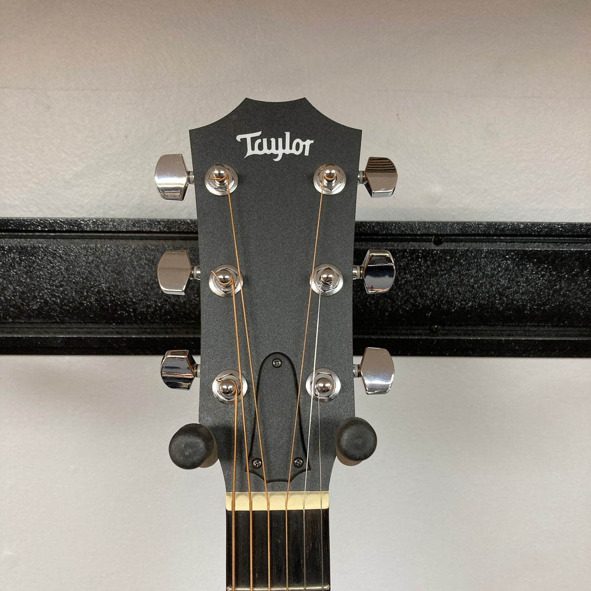 The headstock of a Taylor Academy 10e Acoustic-Electric Guitar w/GigBag with six tuning pegs, displayed on a wall-mounted holder.