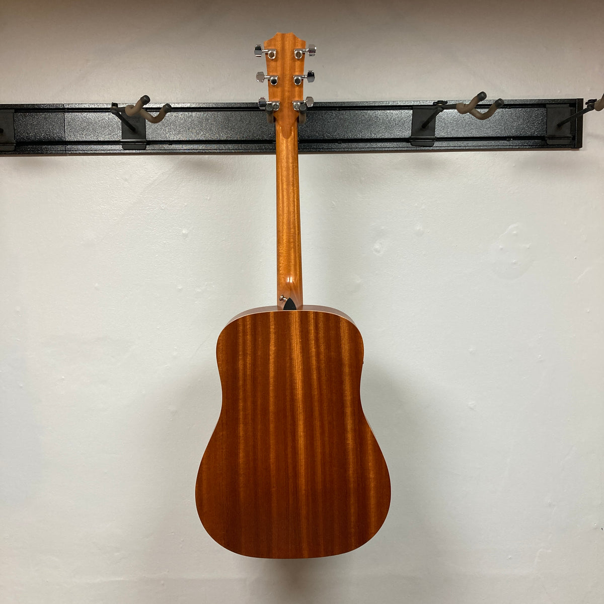 A Taylor Academy 10e Acoustic-Electric Guitar w/GigBag hangs on a black wall rack, showcasing its back and neck against a plain white wall.
