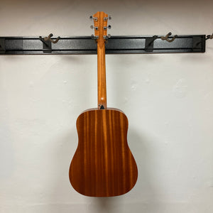 A Taylor Academy 10e Acoustic-Electric Guitar w/GigBag hangs on a black wall rack, showcasing its back and neck against a plain white wall.