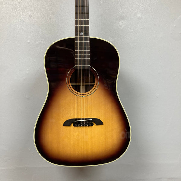 An Alvarez DYMR70SB Yairi Masterworks Acoustic Guitar Sunburst w/Case hangs on a light gray wall, showcasing its stunning sunburst finish.