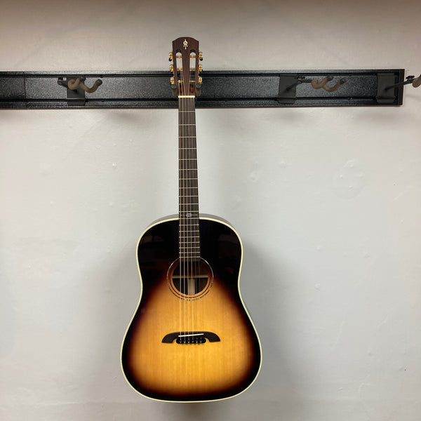 An Alvarez DYMR70SB Yairi Masterworks Acoustic Guitar Sunburst w/Case hangs on a light gray wall, showcasing its stunning sunburst finish.
