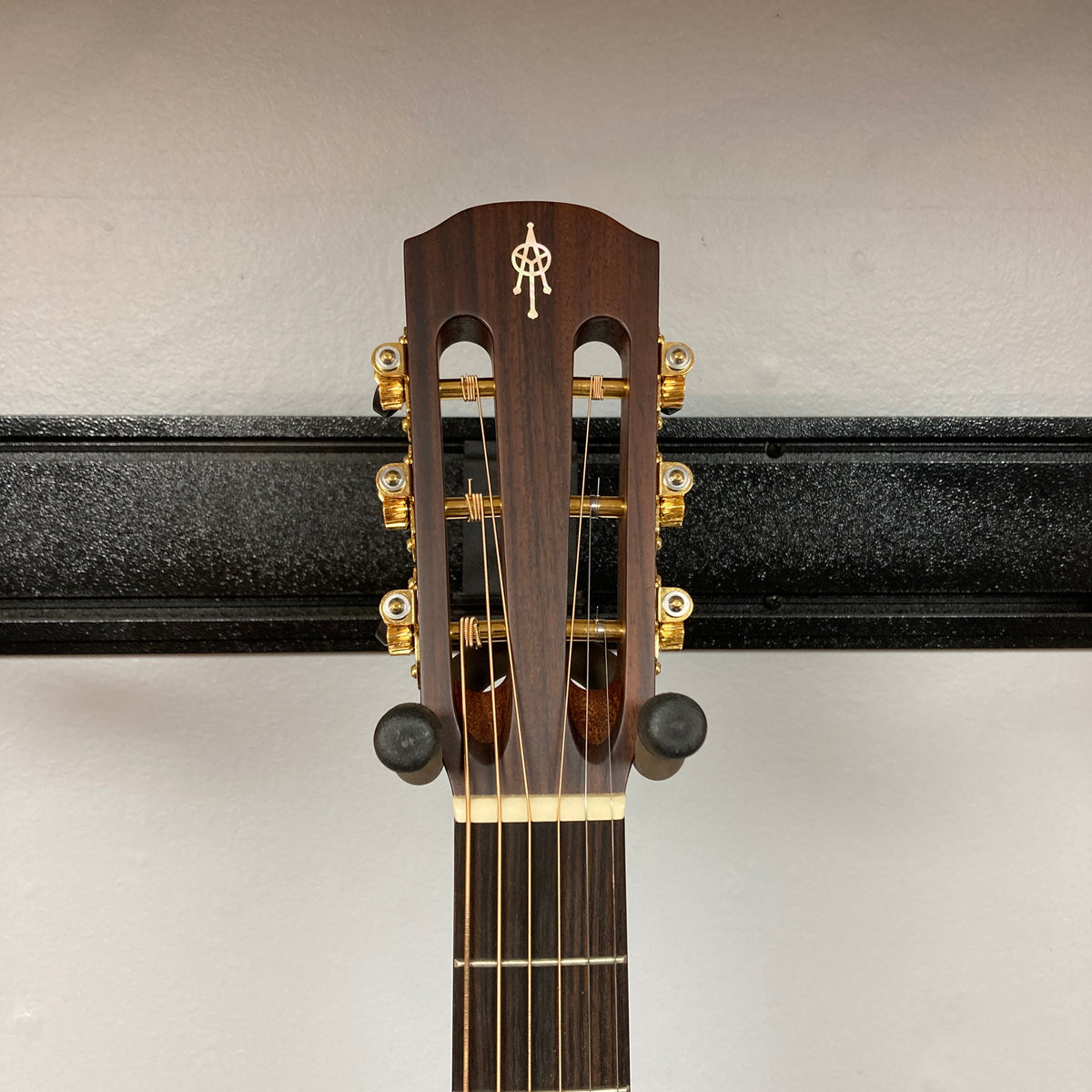 The headstock of an Alvarez DYMR70SB Yairi Masterworks Acoustic Guitar Sunburst w/Case with gold tuners rests on a black wall-mounted guitar holder.
