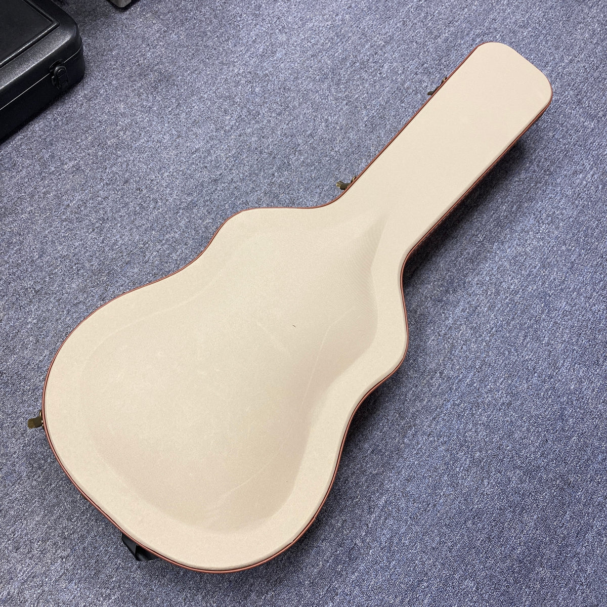A beige guitar case rests closed on a grey carpet, shaped for an Alvarez DYMR70SB Yairi Masterworks Acoustic Guitar Sunburst w/Case inside.