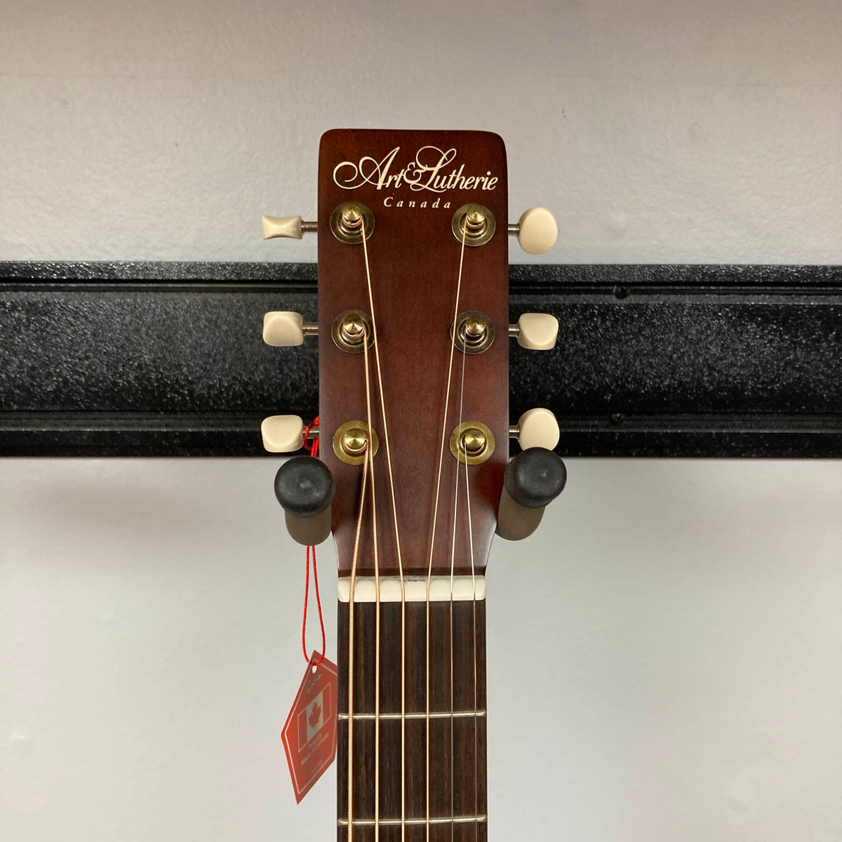 The headstock of the Art & Lutherie Roadhouse Denim Blue Q-Discrete acoustic guitar features tuning pegs, strings, and a red tag, shown against a white wall and black rack.