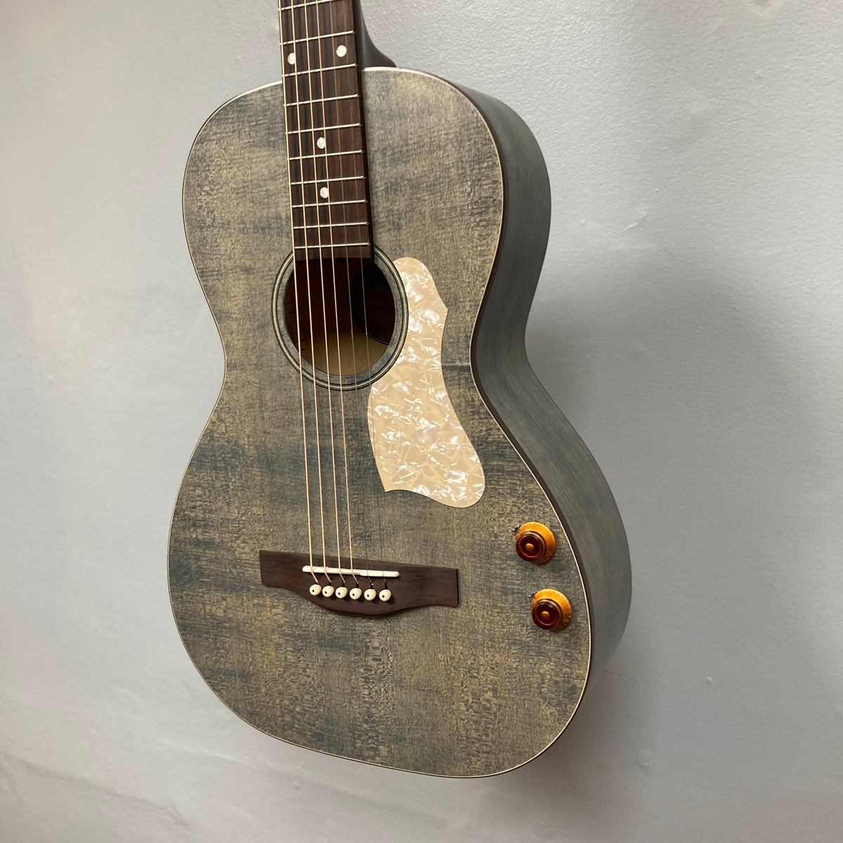An Art & Lutherie Roadhouse Denim Blue Q-Discrete acoustic guitar with six strings, two control knobs, a white pickguard, and a wooden bridge hangs against a plain light-colored wall.