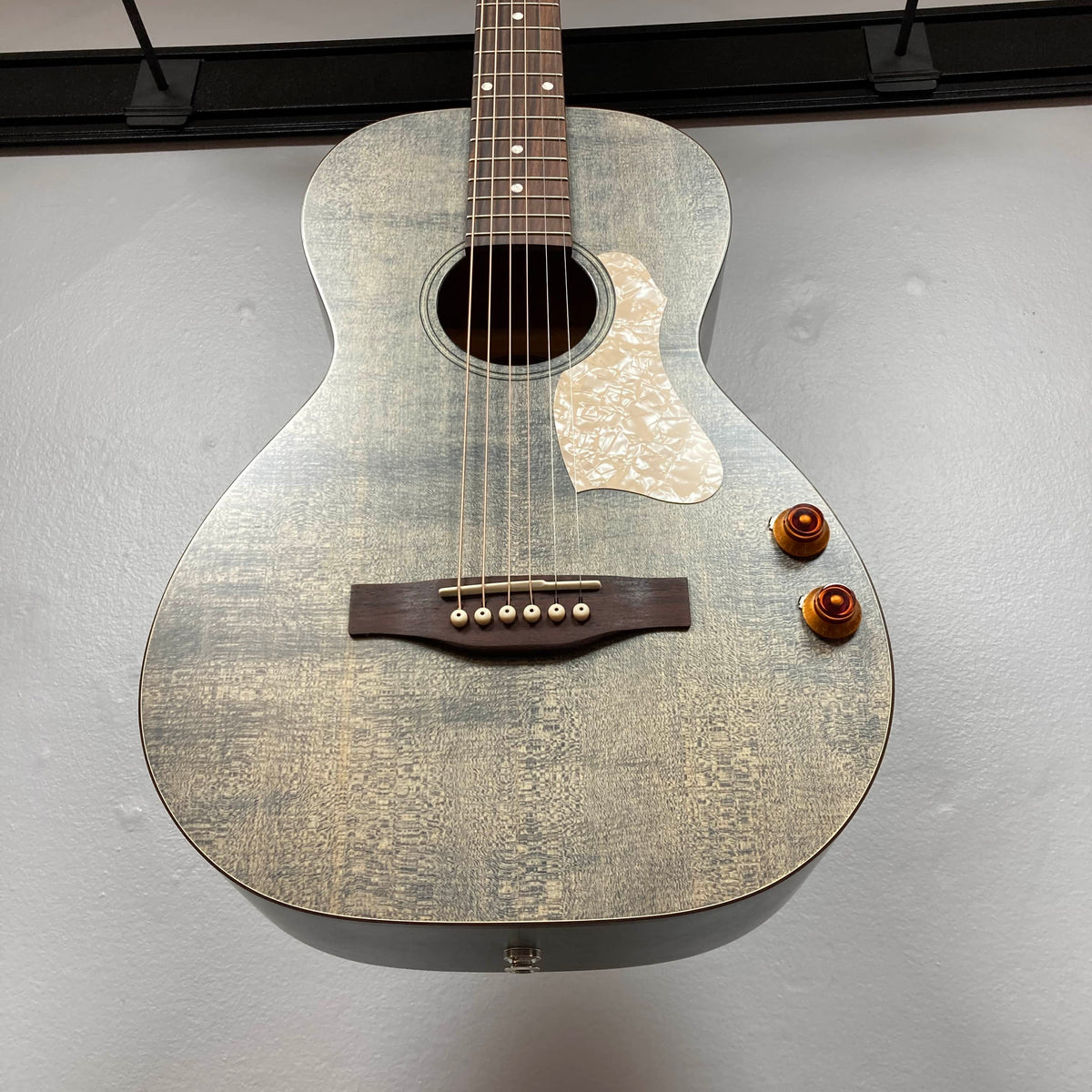 A close-up of the Art & Lutherie Roadhouse Denim Blue Q-Discrete parlor acoustic guitar, featuring a matte finish, two Q-Discrete preamp control knobs, and a decorative pickguard, mounted on a wall.