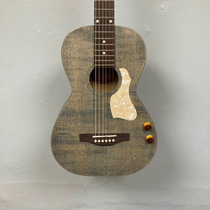 The Art & Lutherie Roadhouse Denim Blue Q-Discrete, a parlor acoustic guitar with a white pickguard, two control knobs, and six strings, hangs against a light gray wall.