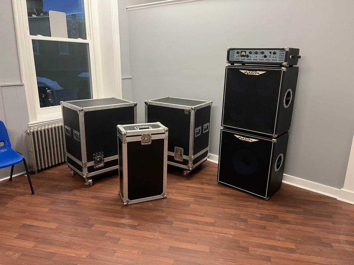 An Ashdown UK Built ABM Bass Rig Full Stack with EVO II 900 Head, 410T and 115-500 cabinets, plus flight cases, rests on a wooden floor in a room with gray walls and a blue chair.