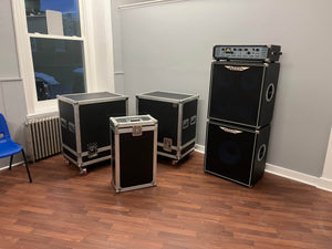 An Ashdown UK Built ABM Bass Rig Full Stack with EVO II 900 Head, 410T and 115-500 cabinets, plus flight cases, rests on a wooden floor in a room with gray walls and a blue chair.