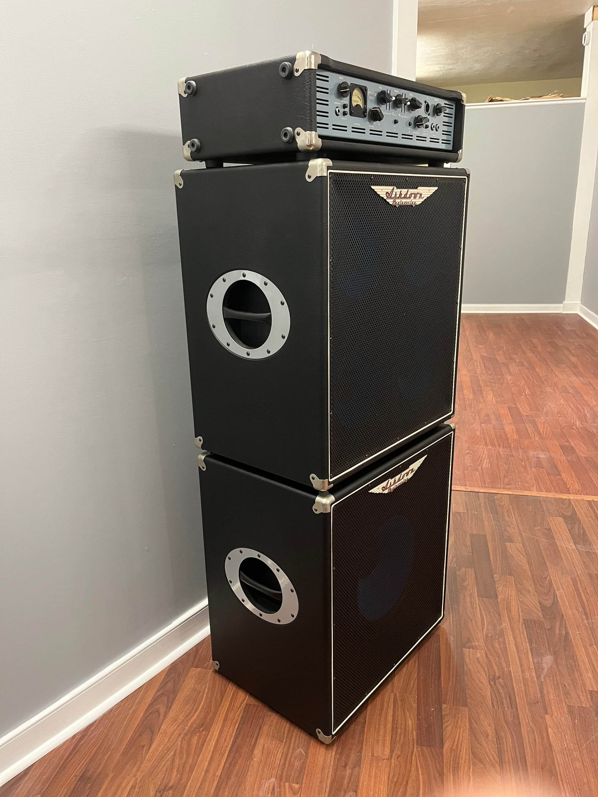 An Ashdown UK Built ABM Bass Rig Full Stack—featuring the EVO II 900 Head atop 410T and 115-500 cabinets with flight cases—creates a powerful setup against a gray wall on a wood floor.