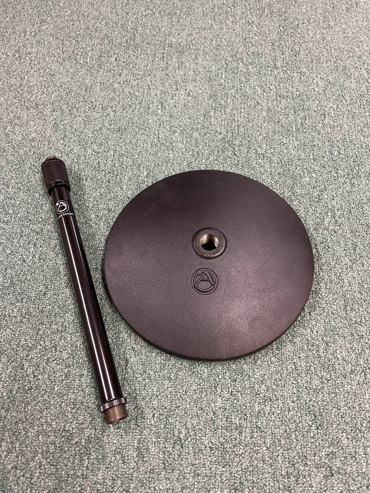 An Atlas Sound DMS10E Mic Stand Base with Extension Rod, both in black and featuring the brand’s logo, are set on a textured green surface.