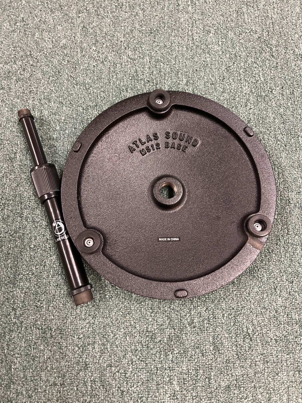 The Atlas Sound DMS7E Mic Stand Base with Extension Rod is shown in black, featuring a round base with three screws and a detachable rod, all placed on a textured green carpet.