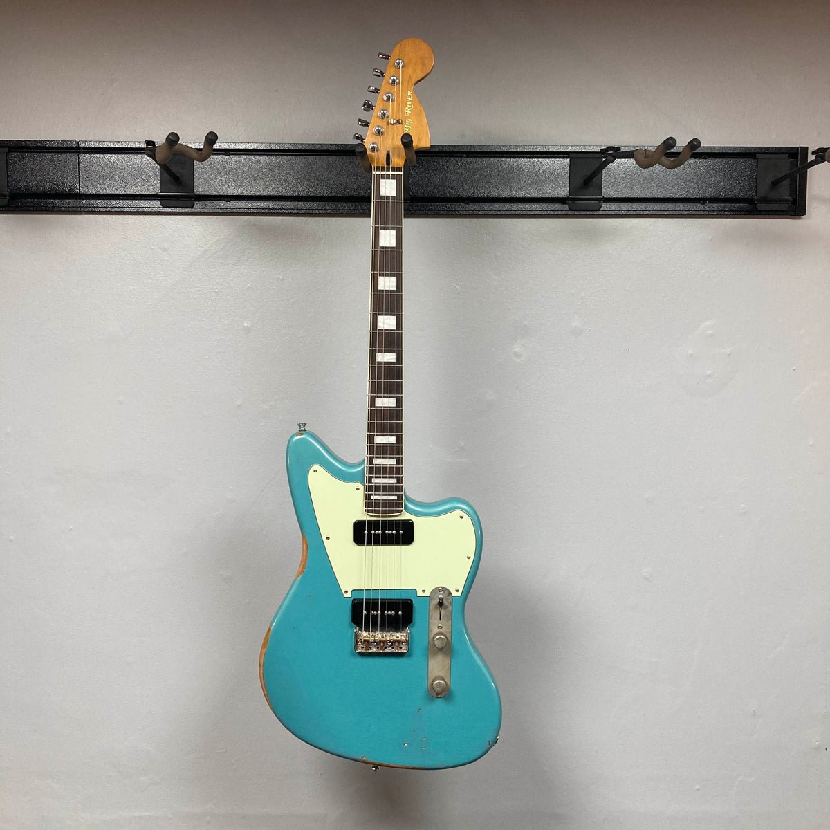 Big River Jazzcaster Custom Relic Pelham Blue electric guitar with a cream pickguard, two Lollar soapbar pickups, and rectangular fret markers, hanging on a wall-mounted rack.