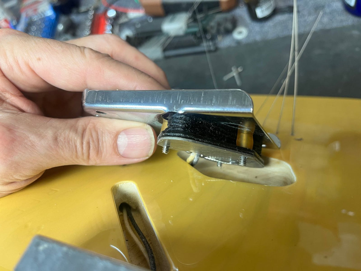 A hand holds a Fender Custom Shop '51 Reissue Nocaster NOS 2006 bridge above a yellow Fender guitar body, with loose wires and components in the background, showcasing vintage Telecaster craftsmanship.