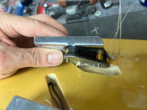 A hand holds a Fender Custom Shop '51 Reissue Nocaster NOS 2006 bridge above a yellow Fender guitar body, with loose wires and components in the background, showcasing vintage Telecaster craftsmanship.