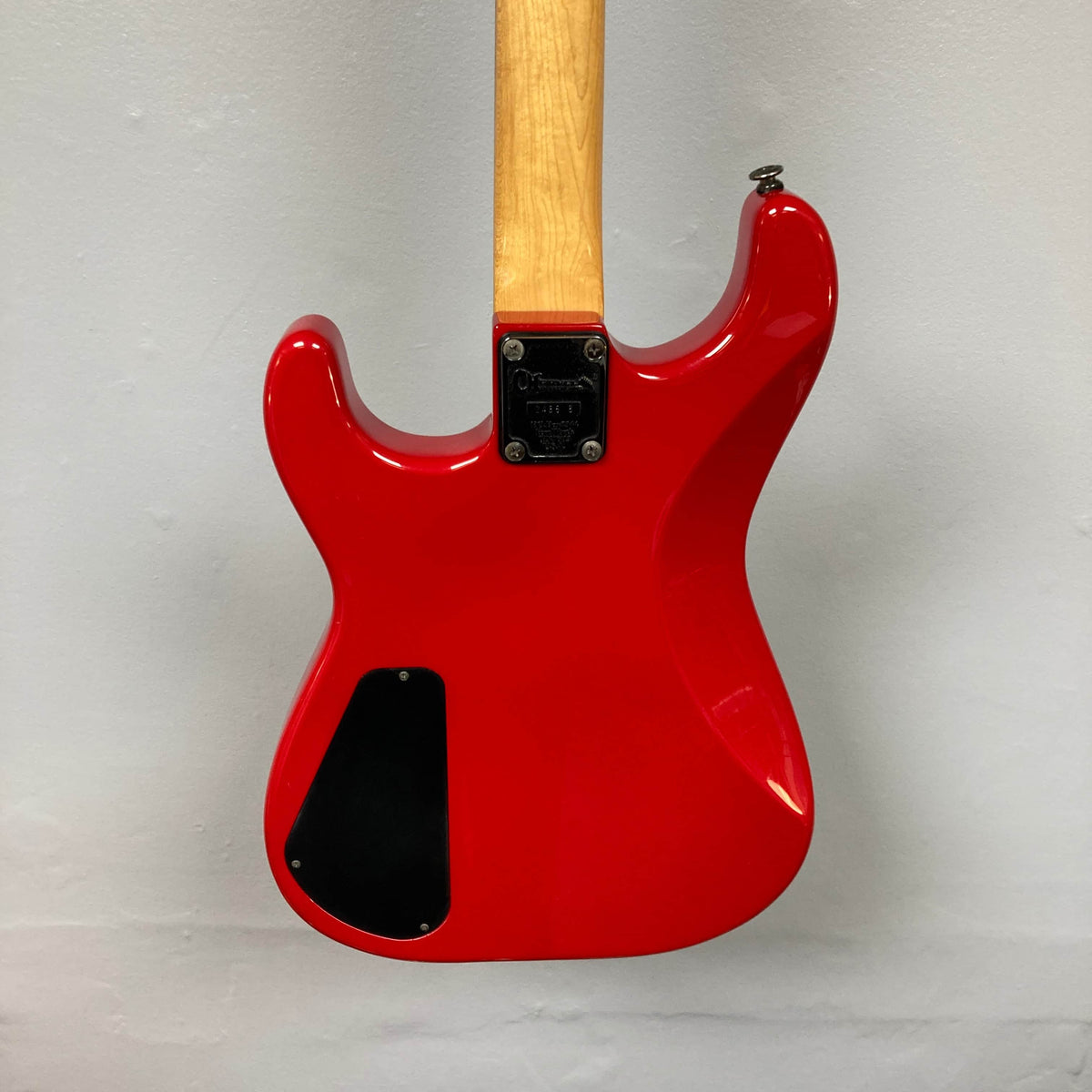 The back of a red Charvel Model 4 HSS electric guitar with a light wood neck, black cover plate, and metal neck plate is shown against a light gray wall. Brand: Charvel. Product: Charvel Model 4 HSS Red w/OHSC Consign.