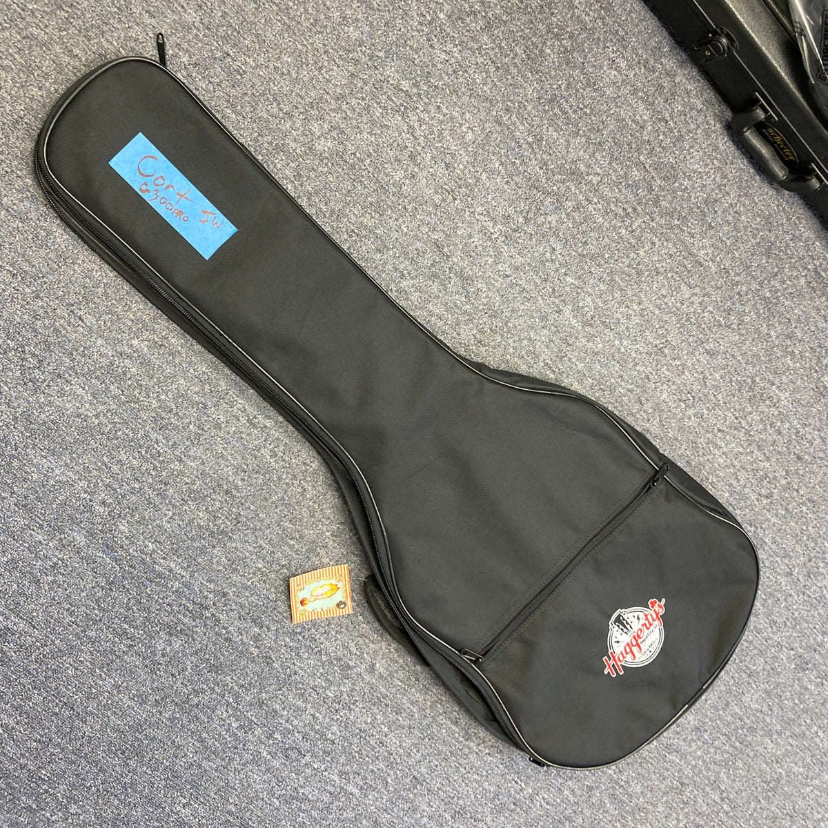 A vivid burgundy Cort FW-G300 PRO guitar with gig bag rests on a gray carpet next to a set of Ernie Ball Regular Slinky guitar strings and a blue label reading "Cort G200 SE.