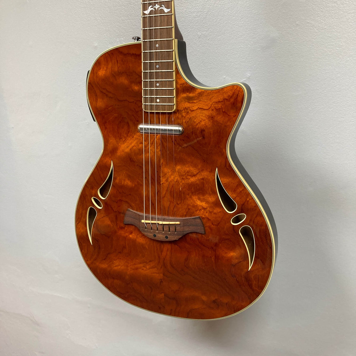 A Crafter SA-BUB Acoustic Electric Guitar Used Consign with a glossy figured wood top, white binding, decorative sound holes, and cutaway design hangs on a light wall—ideal for acoustic or plugged-in playing.