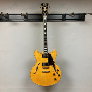 D'Angelico Excel DC Semi-hollowbody Vintage Natural guitar hanging on a wall hook, showcasing double-cutaway design and semi-hollow construction.