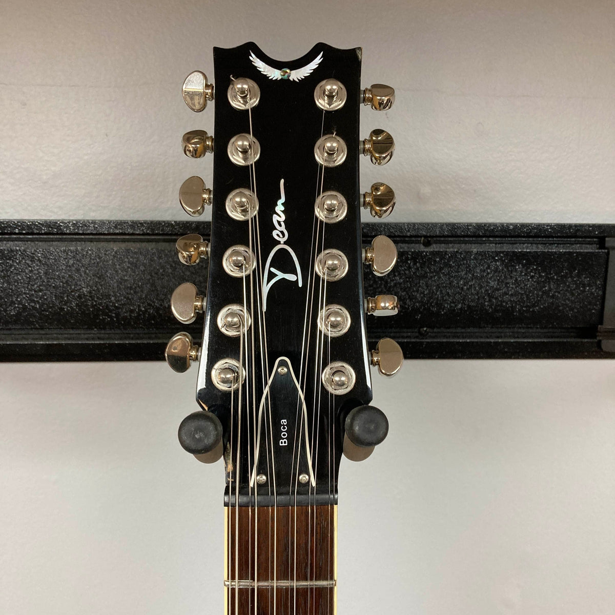 Close-up of the headstock of a Dean Boca Trans Amber Burst 12 String w/Gigbag electric guitar by Dean, mounted on a wall hanger, showing tuning pegs and the Dean logo.