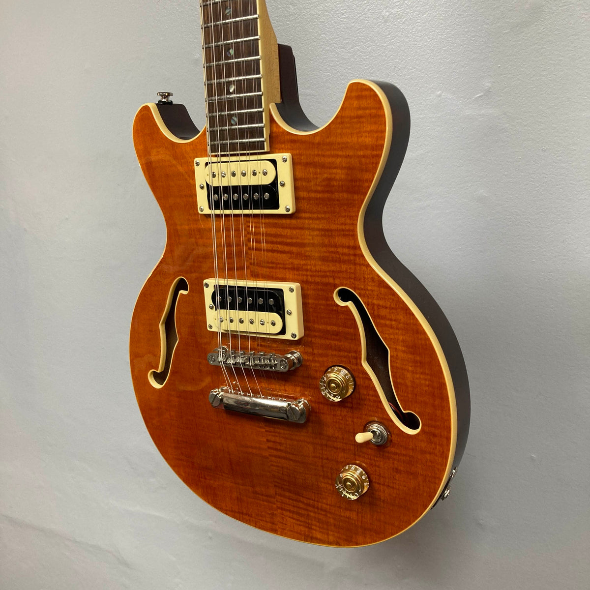 A Dean Boca Trans Amber Burst 12 String guitar by Dean, featuring gold control knobs, f-holes, and a semi-hollow body, is photographed against a plain light gray wall. Comes with gigbag.