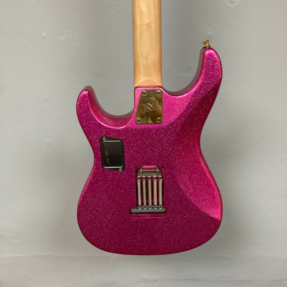 A close-up view of the back of an Edwards ESP E-SN-185-TO Takayoshi Ohmura Twinkle Pink electric guitar, featuring dual humbuckers, a maple neck, metal hardware, and a rectangular battery compartment, set against a plain light gray background.