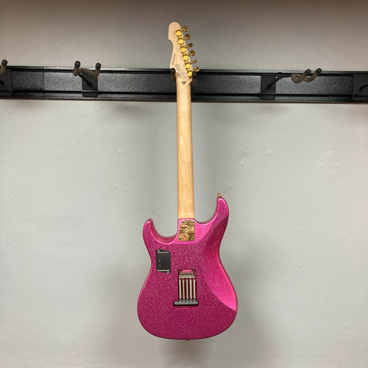 An Edwards ESP E-SN-185-TO Takayoshi Ohmura electric guitar in Twinkle Pink with a glitter finish hangs on a wall-mounted rack, shown from the back to display its neck, tuning pegs, and hardware.