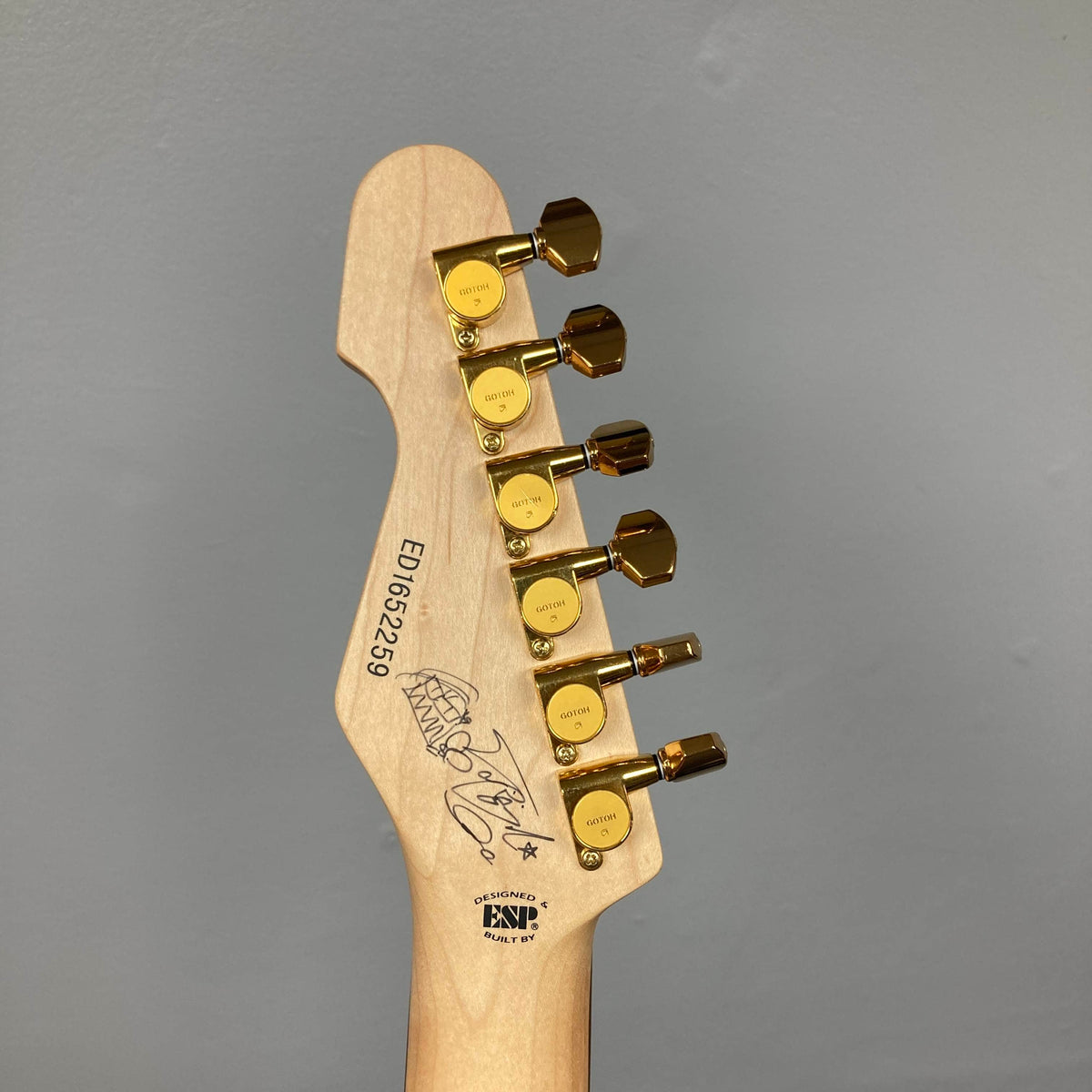 Here's a rewritten version using the provided product data: The back of an Edwards ESP E-SN-185-TO Takayoshi Ohmura Twinkle Pink electric guitar headstock, featuring six gold tuning pegs, serial number, and signature on a light wood finish against a plain gray background.