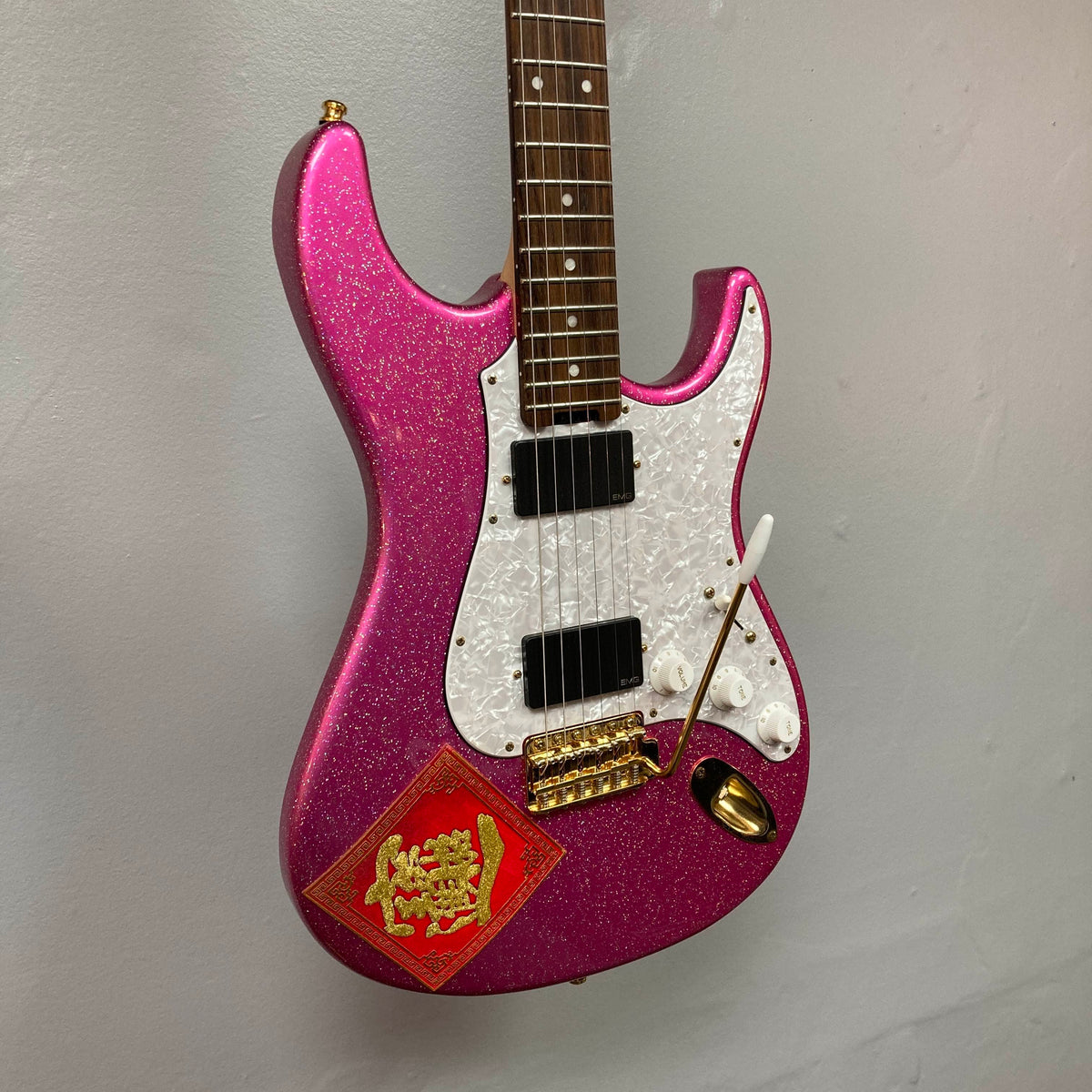 Edwards ESP E-SN-185-TO Takayoshi Ohmura Twinkle Pink electric guitar, featuring dual humbuckers, a sparkling pink glitter finish, white pickguard, gold hardware, and a red decorative sticker in the lower corner, photographed against a plain gray wall.