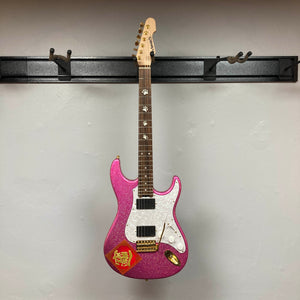 An Edwards ESP E-SN-185-TO Takayoshi Ohmura electric guitar in Twinkle Pink with a white pickguard and a red sticker is hanging on a black wall mount against a plain light-colored wall.