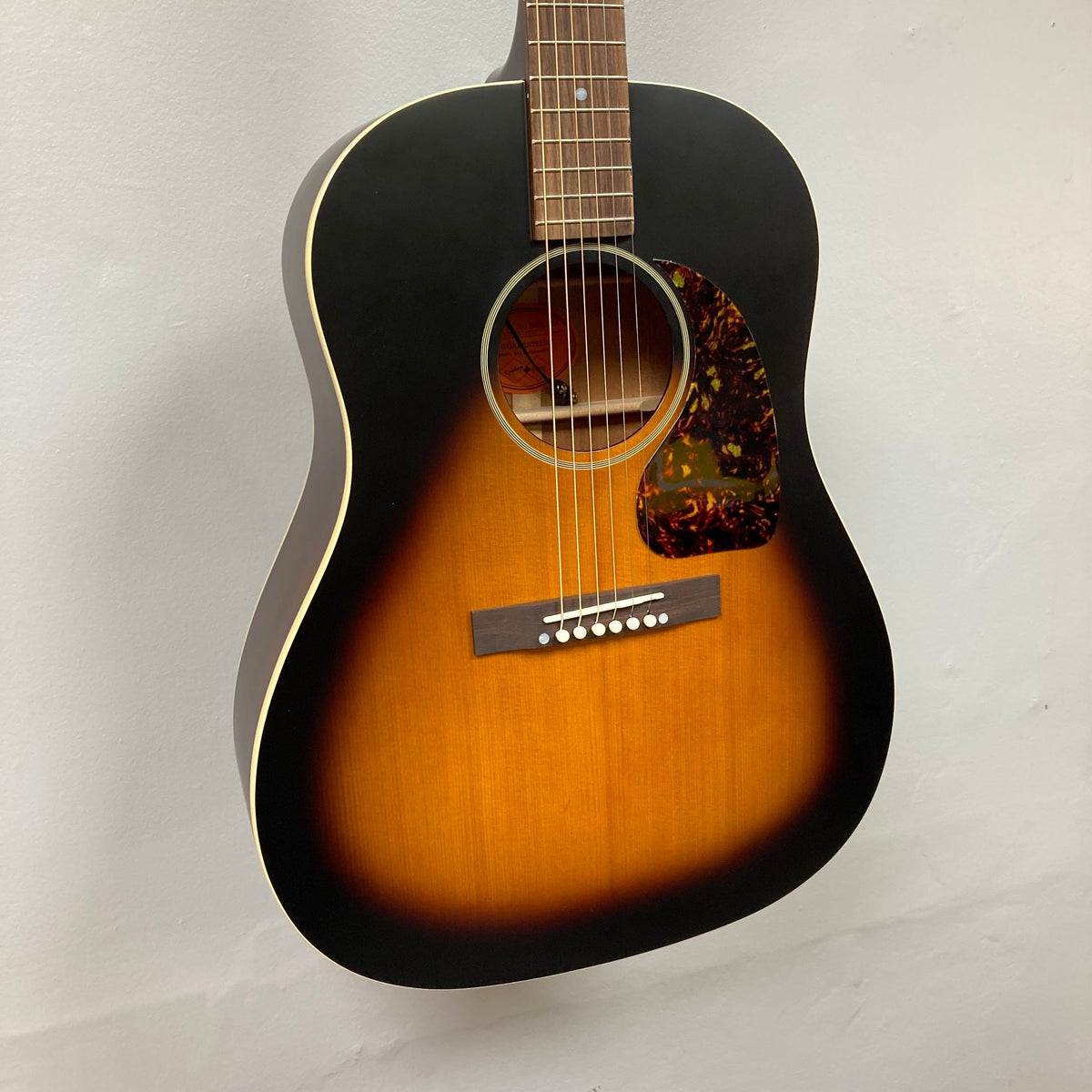 Epiphone 1942 Banner J-45 Vintage Sunburst acoustic guitar with mahogany body, spruce top, and V-shaped neck, displayed on a wall, includes hardshell case.