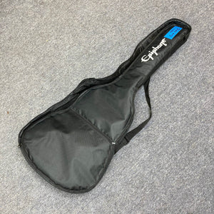 A black Epiphone soft case, ideal for the Epiphone Blues King Studio Dark Burst acoustic guitar, features a shoulder strap and front zipper pocket. It rests on gray carpet with a blue numbered sticker on the headstock area.