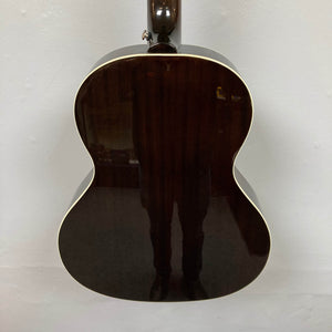 The back of an Epiphone Blues King Studio Dark Burst acoustic guitar, featuring a dark glossy finish and light binding, is shown hanging against a plain wall with a person's reflection visible on its shiny surface.