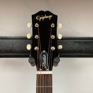 A close-up of the headstock of the Epiphone Blues King Studio Dark Burst acoustic guitar shows cream tuning pegs, a gold Epiphone logo, and “Studio” on the truss rod cover, set against a light-colored wall.
