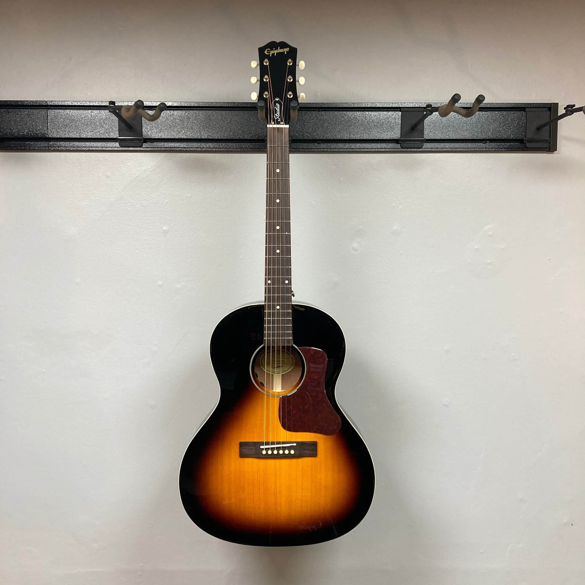 The Epiphone Blues King Studio Dark Burst acoustic guitar by Epiphone, featuring a dark burst finish, hangs on a wall-mounted holder against a plain white wall, highlighting its classic blues folk style.