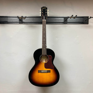 The Epiphone Blues King Studio Dark Burst acoustic guitar by Epiphone, featuring a dark burst finish, hangs on a wall-mounted holder against a plain white wall, highlighting its classic blues folk style.