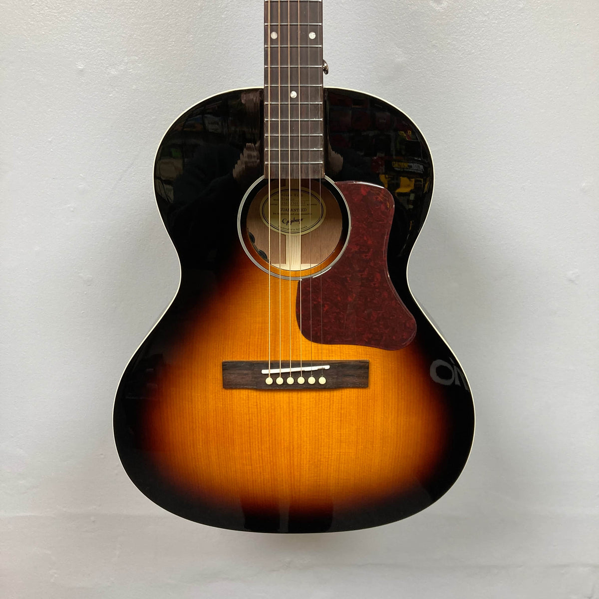 The Epiphone Blues King Studio Dark Burst, a six-string folk guitar with a dark-to-light sunburst finish and tortoiseshell pickguard by Epiphone, hangs vertically against a plain light-colored wall.
