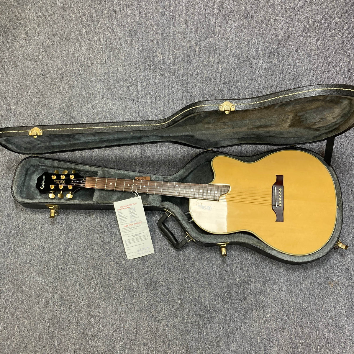 An Epiphone Chet Atkins SST Studio Natural acoustic-electric guitar by Epiphone rests in an open hard case with gold-tone latches and a handle, on a grey carpeted floor. A paper card is placed on the body. Used consignment, made in Korea.