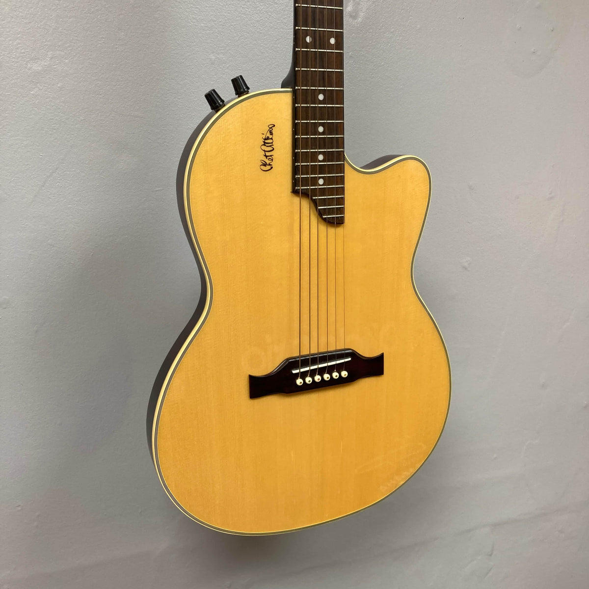 A close-up of an Epiphone Chet Atkins SST Studio Natural acoustic-electric guitar, made in Korea, with a light wood top and cutaway body, mounted on a light gray wall. Includes hard case; used consignment.