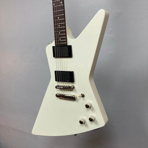 A Classic White Epiphone Explorer 80s EMG electric guitar with sharp, angular design, black pickups, chrome hardware, and three control knobs stands against a plain wall; includes gigbag.