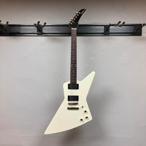 The Epiphone Explorer 80s EMG Classic White electric guitar with a mahogany neck and black headstock hangs on a wall-mounted rack against a light gray wall, evoking classic rock style. Includes gigbag.