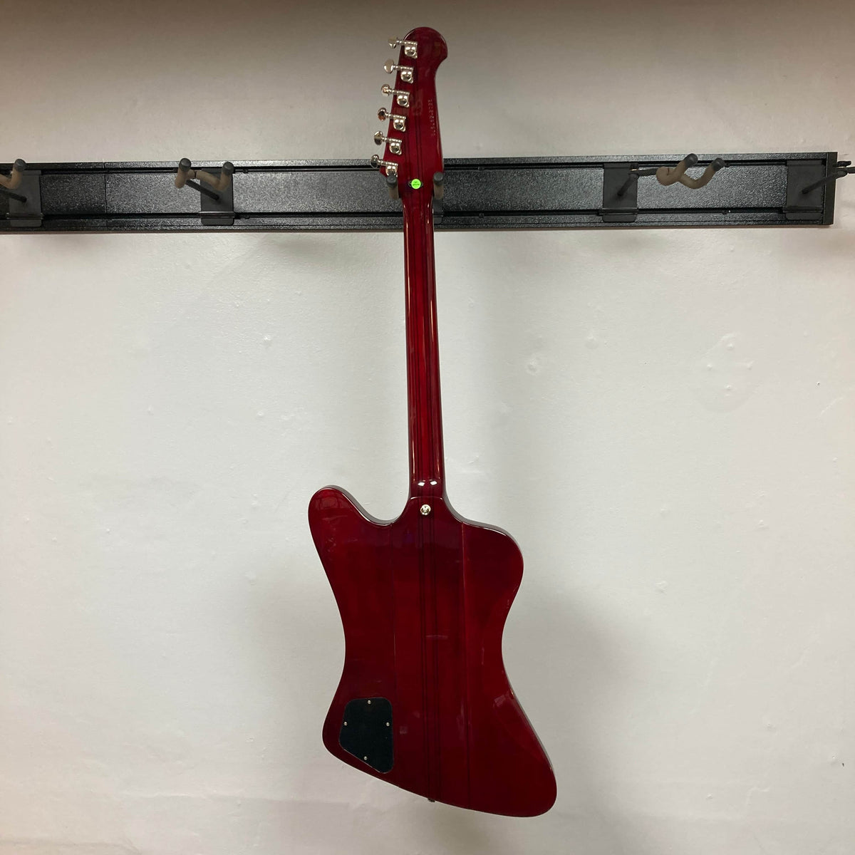 An Epiphone Firebird Wine Red electric guitar with a neck-through-body design hangs on a wall-mounted rack, seen from the back. Comes with gigbag.