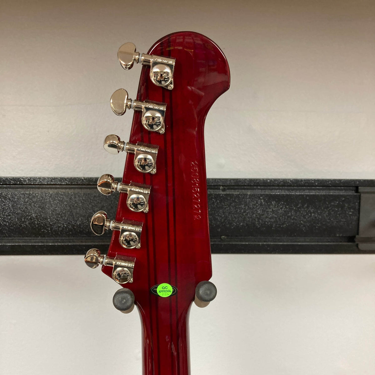 Epiphone Firebird Wine Red w/Gigbag electric guitar by Epiphone features chrome tuning pegs, a serial number on the back of the headstock, a green inspection sticker, and visible neck-through-body design as it hangs on a wall hanger.