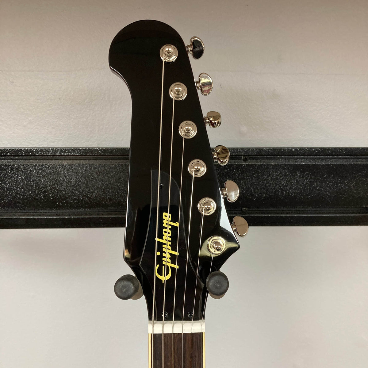 Close-up of the headstock of an Epiphone Firebird Wine Red electric guitar with six tuning pegs and Epiphone logo, displayed on a wall hanger; features a rosewood fretboard that adds warmth and character to its look. Comes with gigbag.