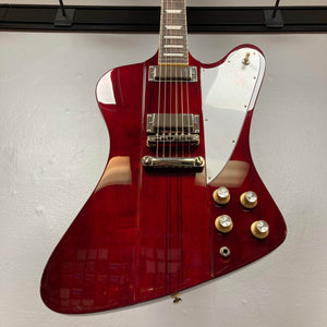 An Epiphone Firebird Wine Red electric guitar with gold hardware, rosewood fretboard, four knobs, and a white pickguard is displayed against a plain light-colored wall. Includes gigbag.