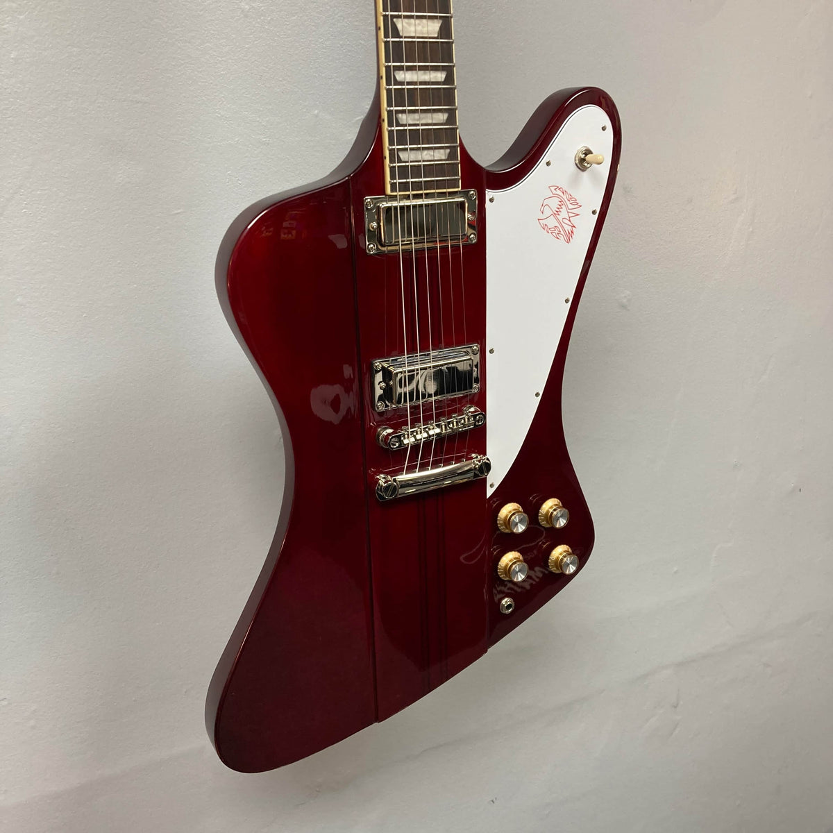 An Epiphone Firebird Wine Red electric guitar with rosewood fretboard, white pickguard, chrome hardware, four control knobs, two pickups, and neck-through-body construction hangs on a white wall.
